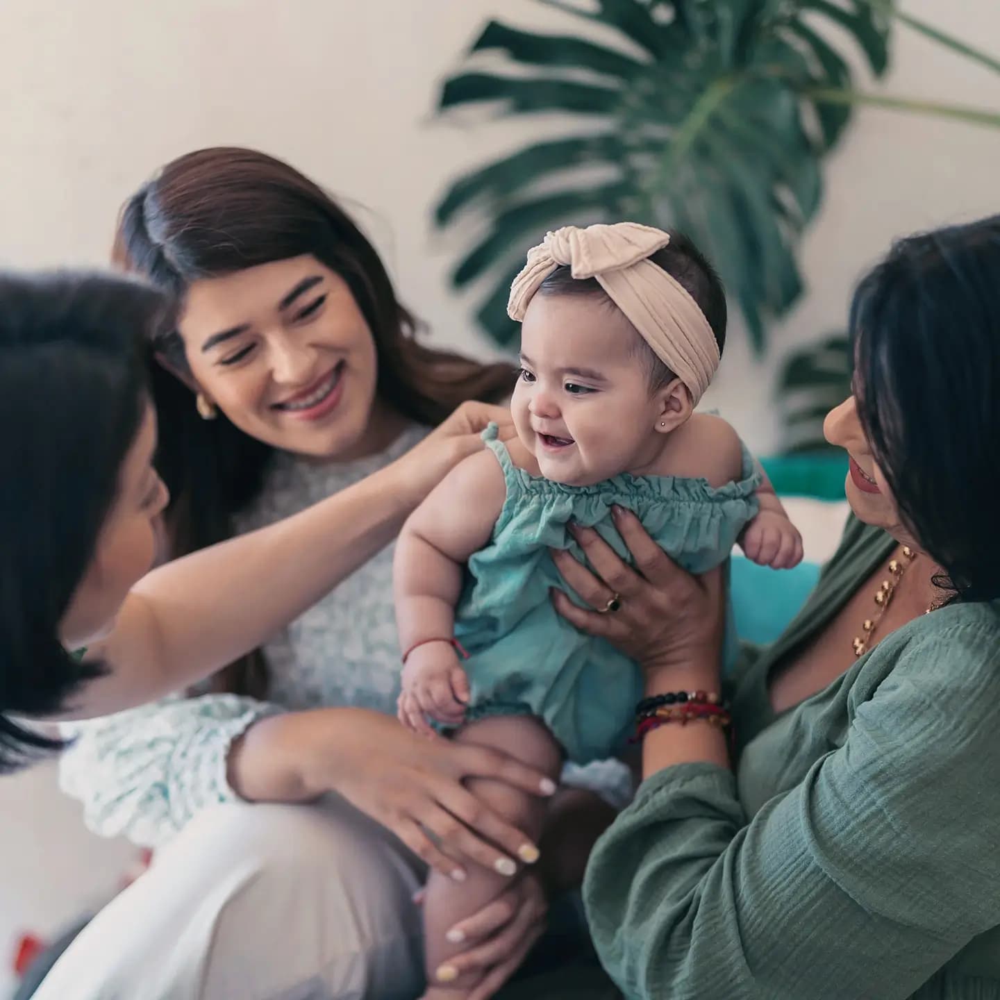 A  baby dressed in teal clothes, surrounded by three smiling women who are all holding and looking lovingly at the baby.