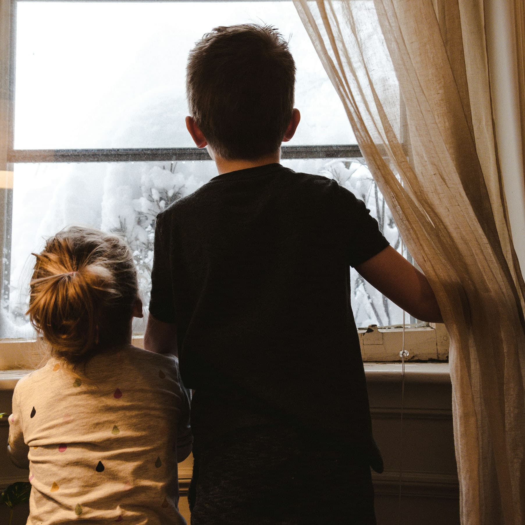 Two little boys, one older than the other, standing together and looking out a window at the snowy scene outside, with the older boy holding a white curtain to the side.