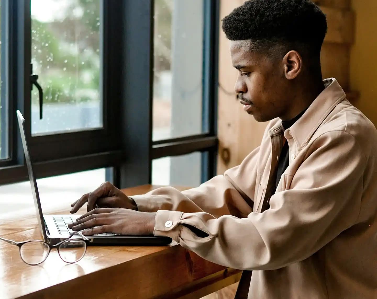 A young man in a light brown jacket focuses on his laptop while sitting at a table in front of a rainy window.