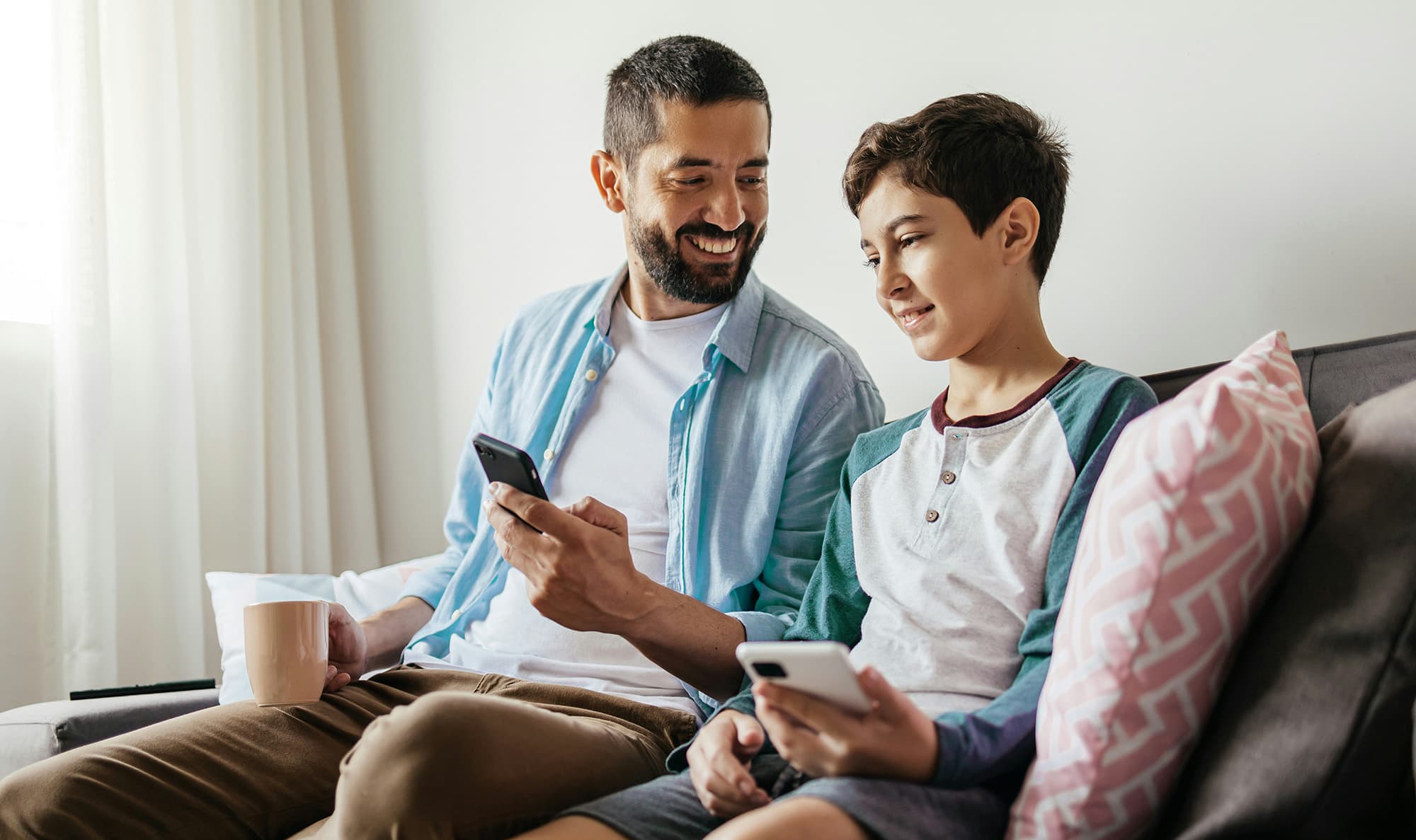 A father and son sit side by side on a sofa; the bearded man, wearing a T-shirt with a light blue shirt over it, smiles while holding a mug in one hand and showing his smartphone to his son, who smiles back while holding his own phone.