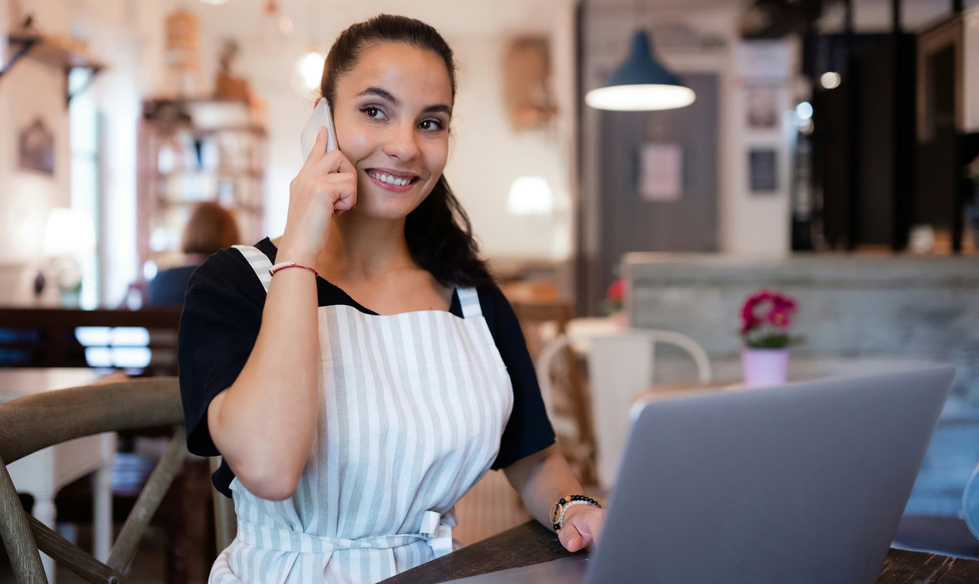 A person wearing a striped apron over a black T-shirt sits at a café table, smiling while talking on the phone, with an open laptop in front of them.
