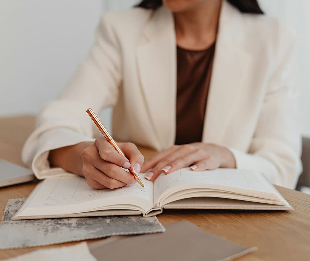 A woman in a white suit takes notes at a wooden desk using an orange pen, with the focus on the open notebook in front of her.