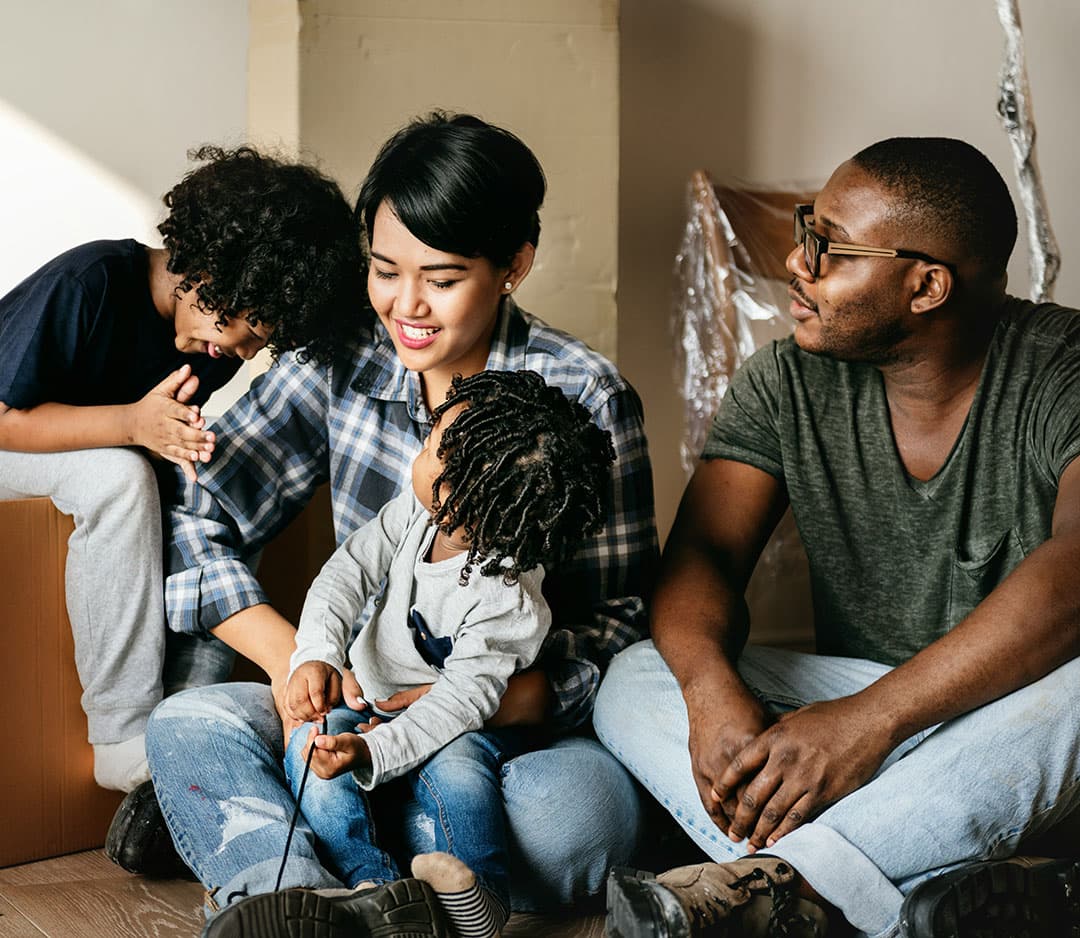 A family sits together with two laughing kids beside their mother, who wears a plaid shirt, while the father sits next to them watching warmly. 