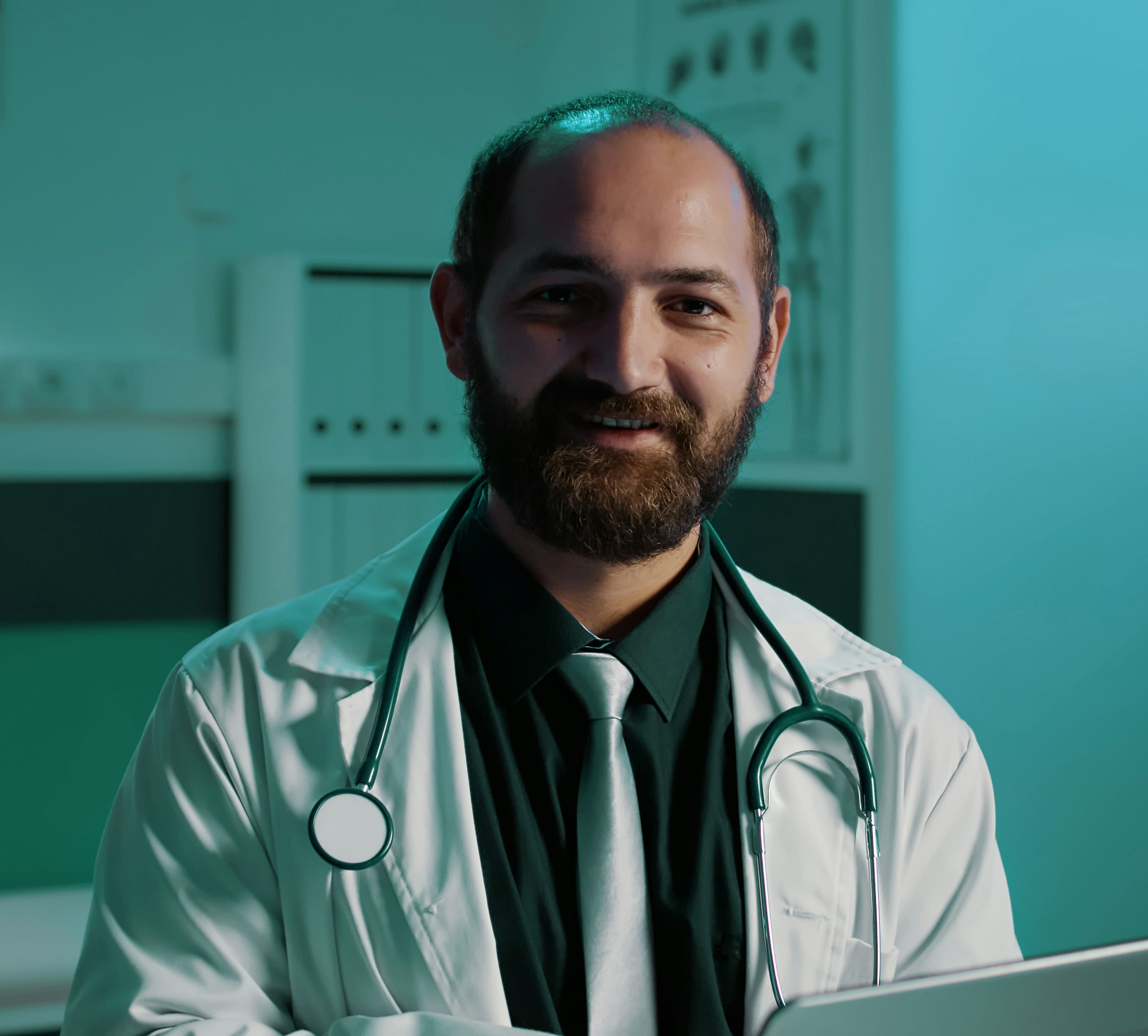 A bearded male doctor stands in a teal-lit room, wearing a lab coat over a black shirt and teal tie, with a stethoscope around his neck.