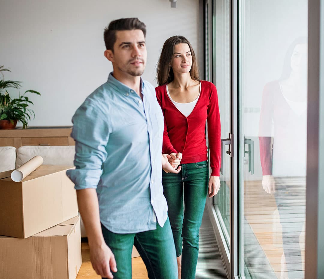 A man in a light blue top and teal jeans leads a woman wearing a red cardigan and teal jeans as they both look out a floor-to-ceiling window; their home is filled with cardboard boxes, suggesting they’ve just moved in.