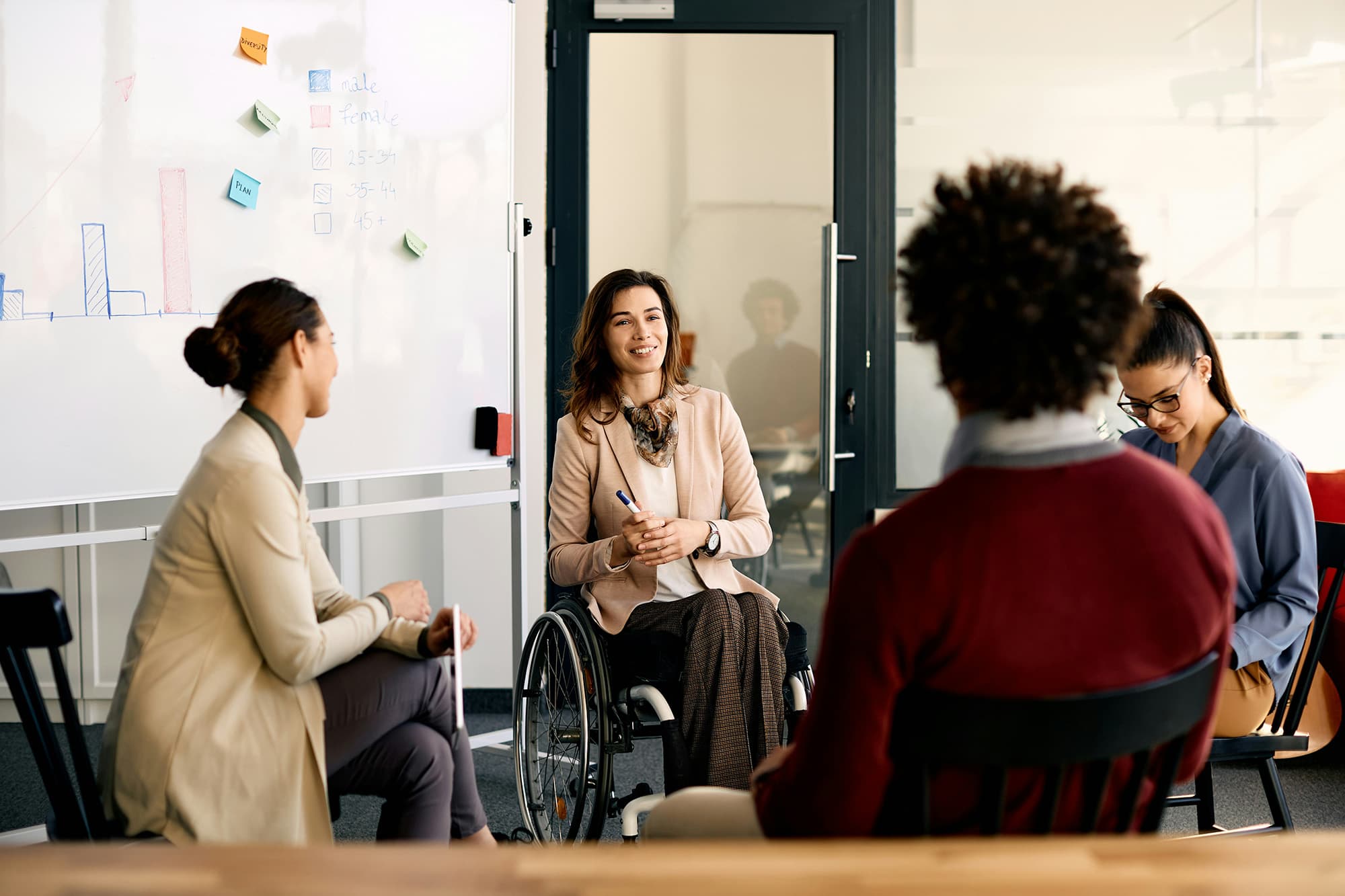 A woman in professional attire sits in a wheelchair beside a whiteboard, speaking as others seated around her listen attentively.