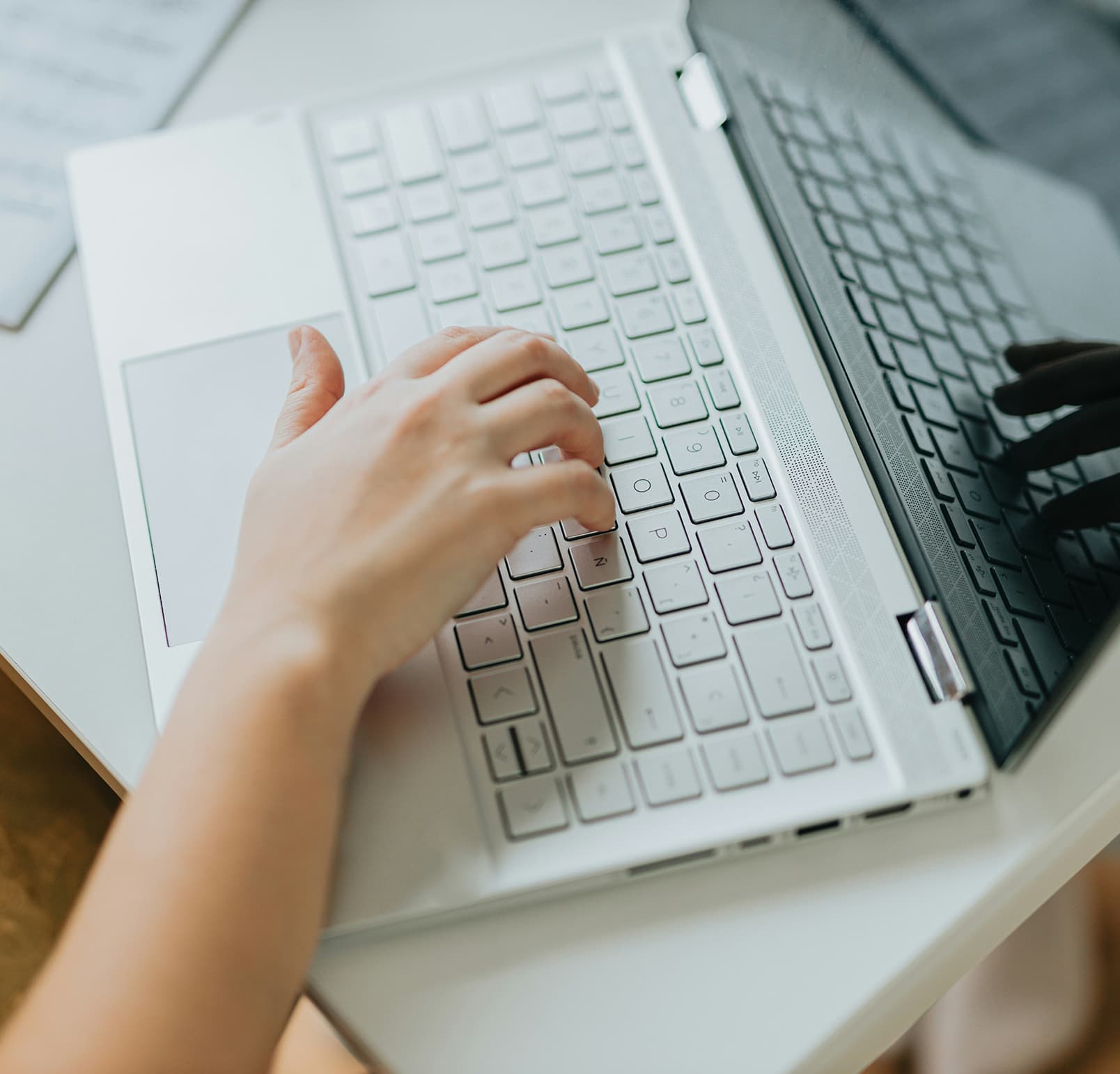 A close-up of a hand typing on the keyboard of a silver laptop placed on a white desk.