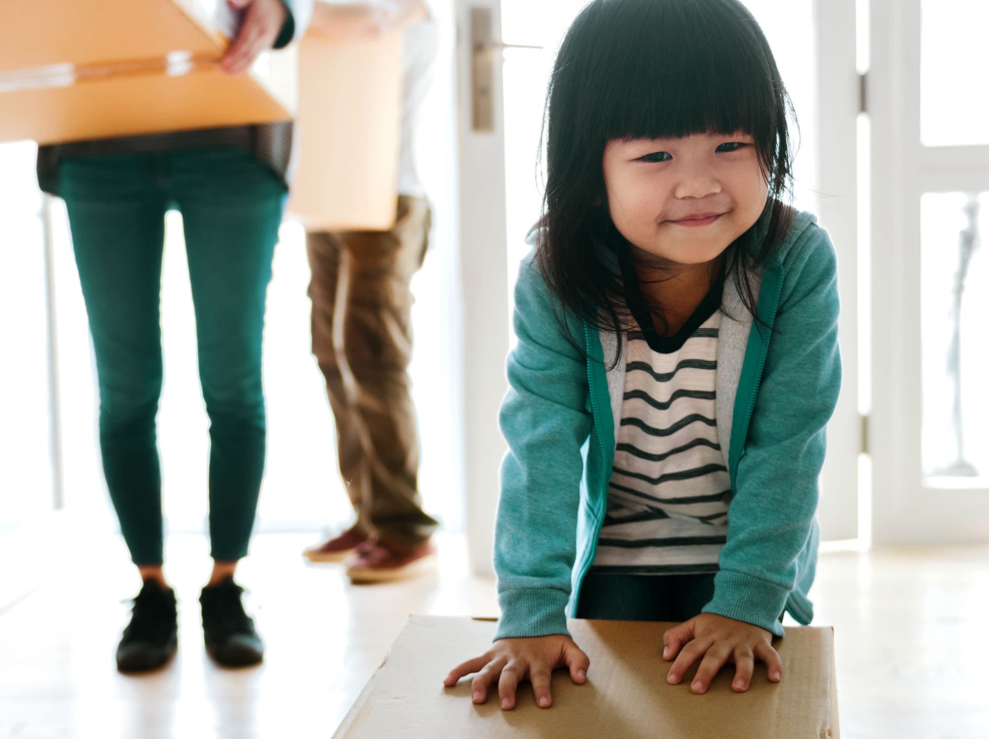 A young girl with bangs wearing a teal jacket places her hands on a cardboard box and pushes it forward, while in the background, her parents’ legs are visible as they carry boxes, suggesting the family is moving in.