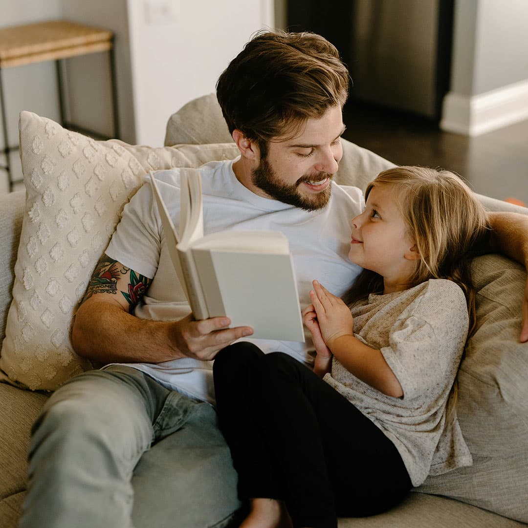 A father with a white t-shirt and a tattoo on his arm and his young daughter sit on a white couch with an off-white cushion, smiling at each other while reading. 