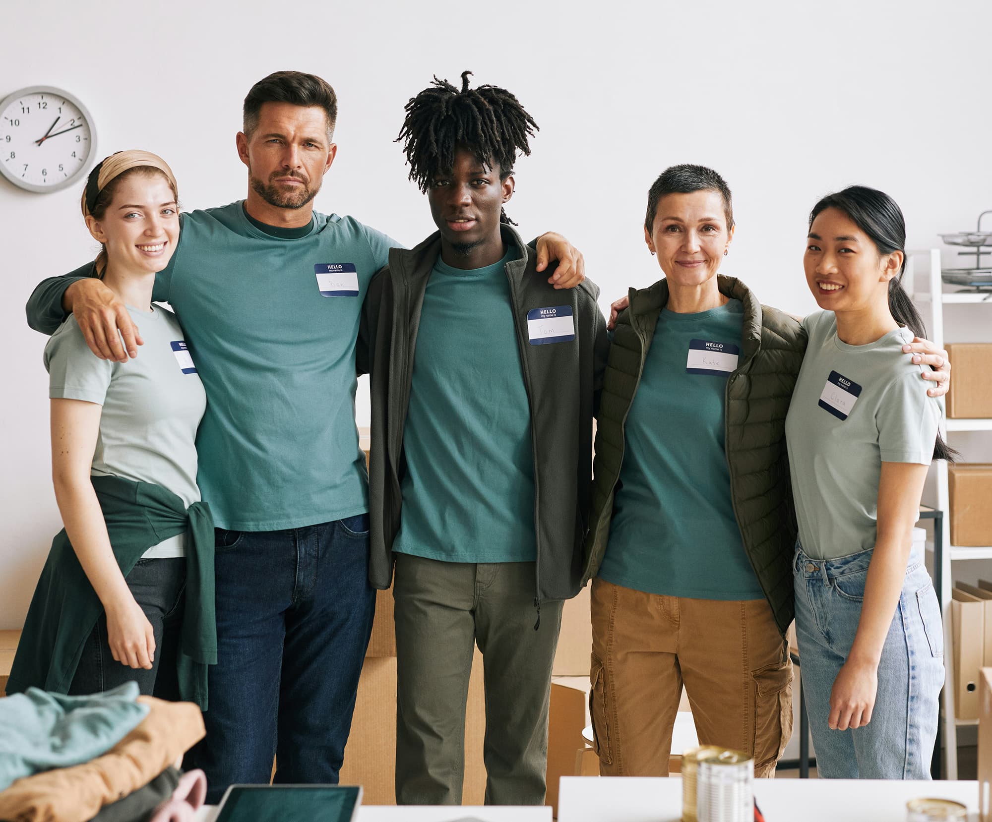 A group of five volunteers wearing varied shades of teal-colored clothing and name tags stand together in a room filled with cardboard boxes.