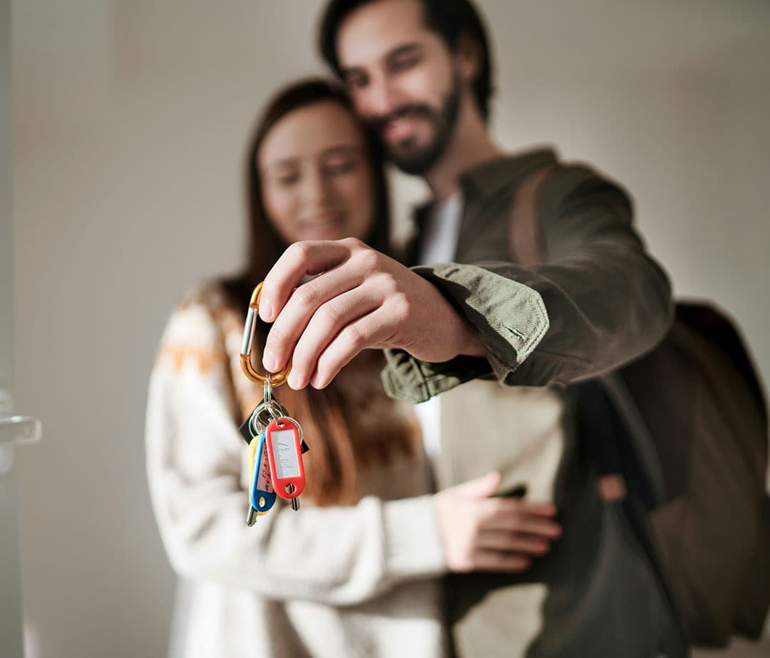 A close-up focused on a set of keys being held by a couple who are blurred in the background as they look at the keys.