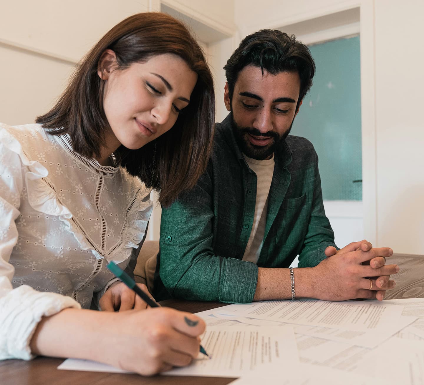 A woman and a man sit close together as the woman signs a document with a pen, while the man watches attentively beside her.
