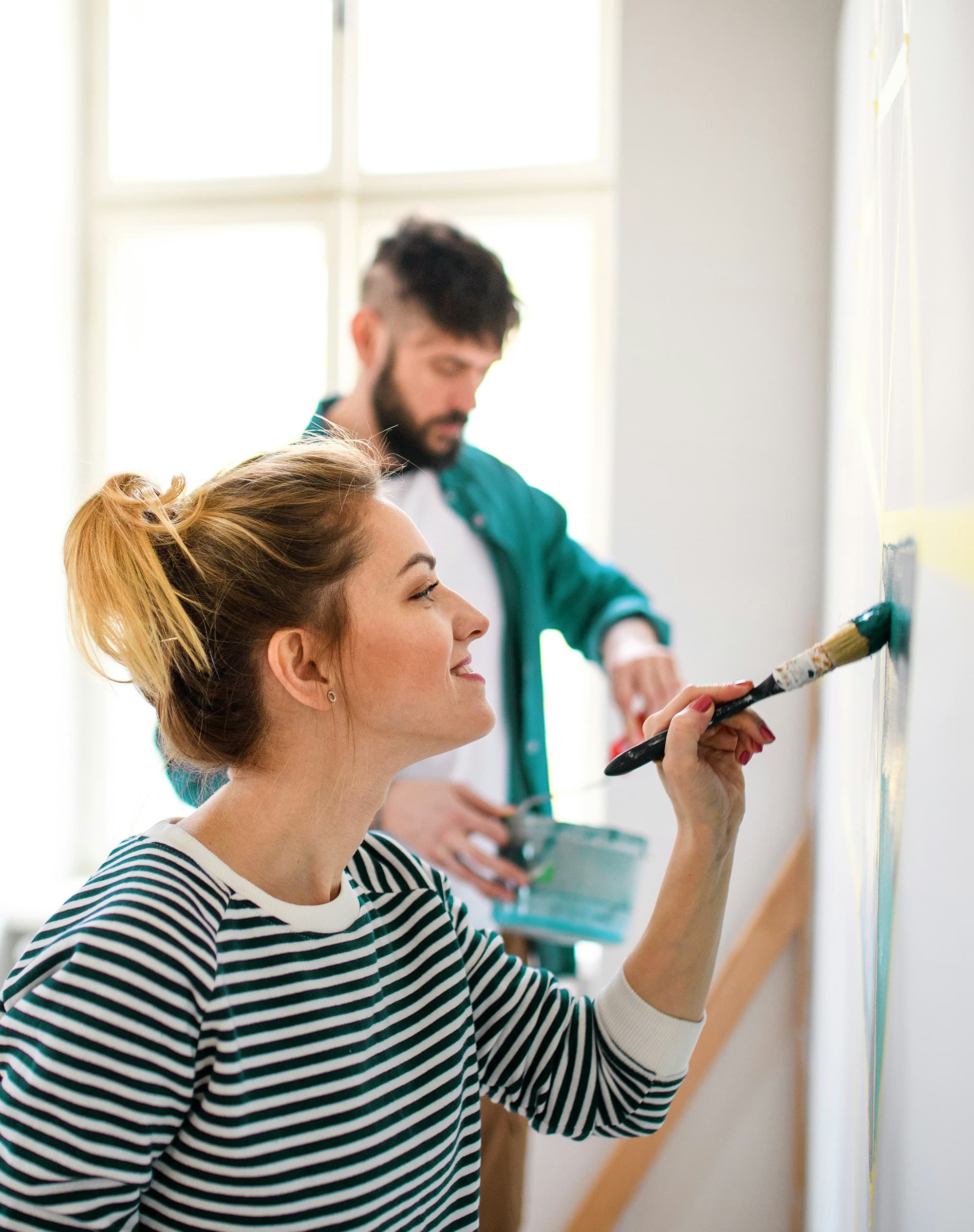 A close-up of a blonde woman painting a white wall with teal paint, wearing a striped shirt, while in the blurred background, a man in a teal shirt layered over a white shirt is visible.
