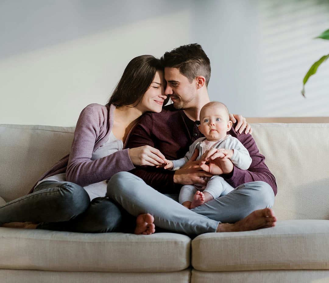 A family sits together on a white couch; a man in a red wine-colored top holds a baby, while a woman hugs him from the side, smiling warmly.