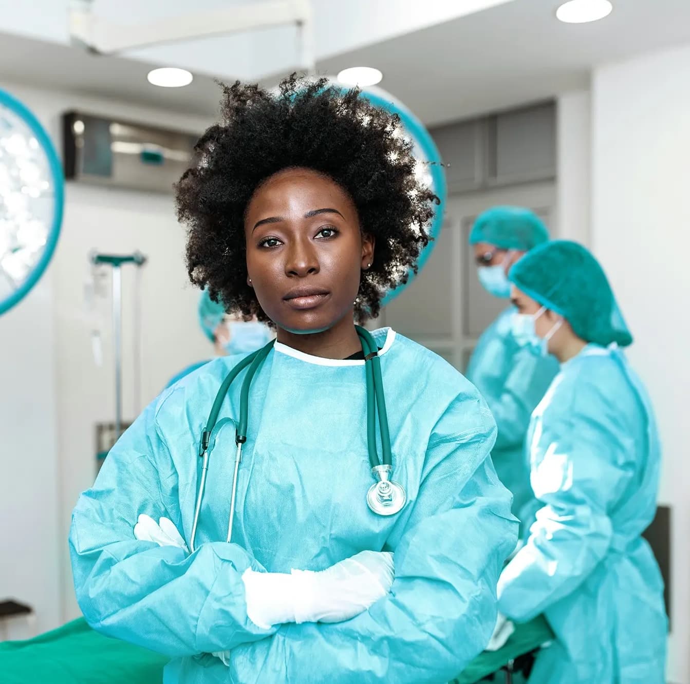 A female surgeon stands confidently in a surgical room, wearing a light blue plastic suit and plastic gloves, with a teal stethoscope hanging around her neck and her arms crossed, while colleagues in similar attire work in the blurred background.