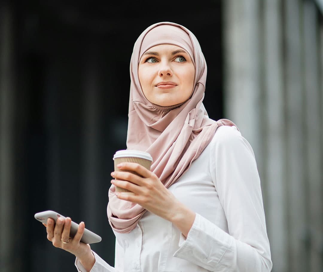 A woman wearing a white shirt and a pink hijab holds an iPhone in one hand and a coffee cup in the other, looking to the side.