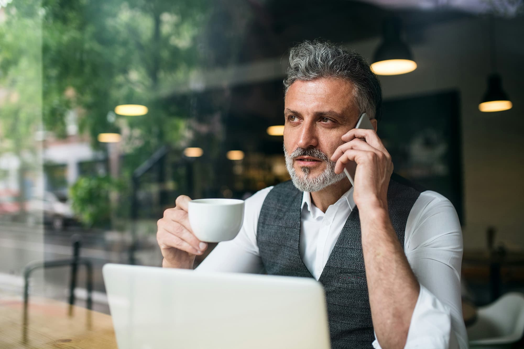 A man in a café is seen through a glass window, holding a phone to his ear and talking while his other hand holds a coffee mug; a laptop sits open in front of him as he looks outside.