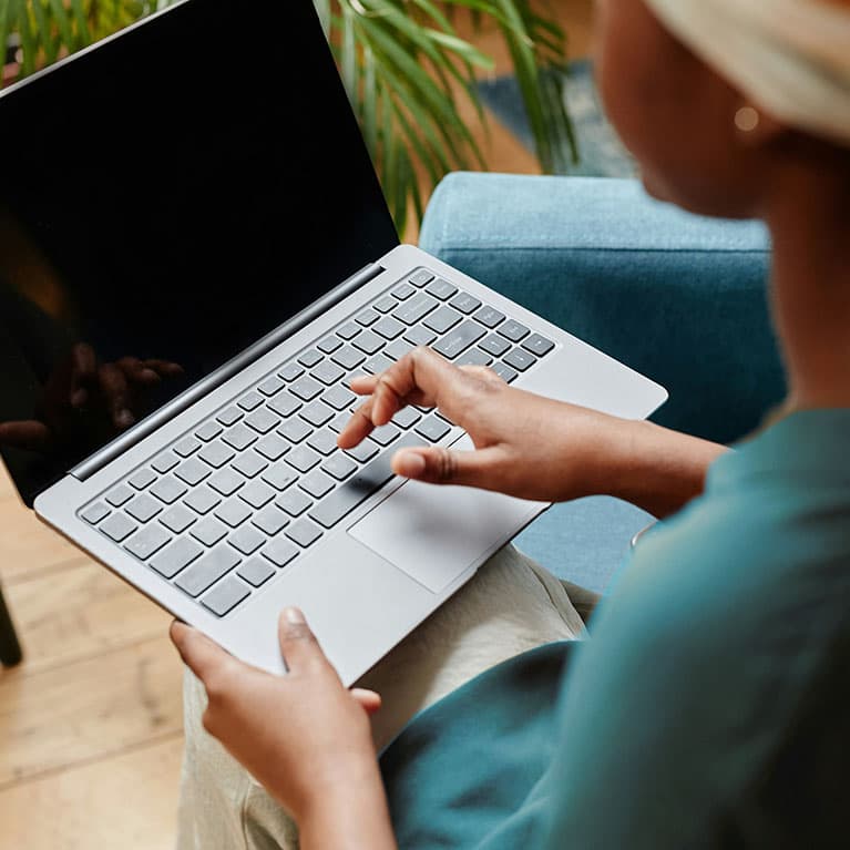 A close-up of one hand typing on a laptop keyboard while the other hand holds the silver computer; the person is wearing a teal top and sitting on a blue sofa.