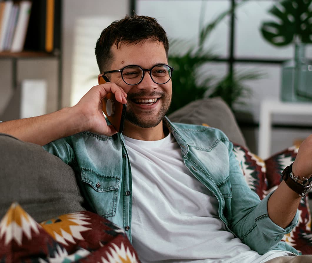A man wearing glasses and a teal jean jacket over a white T-shirt sits on a sofa, smiling as he talks on the phone.