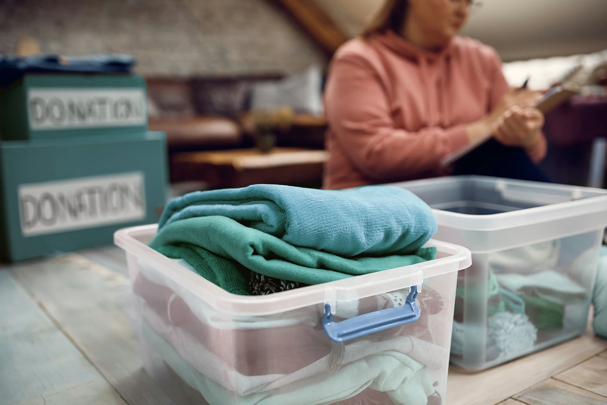 A close-up of teal clothes neatly folded and placed in semi-transparent containers on a table, with a woman writing on a clipboard blurred in the background.