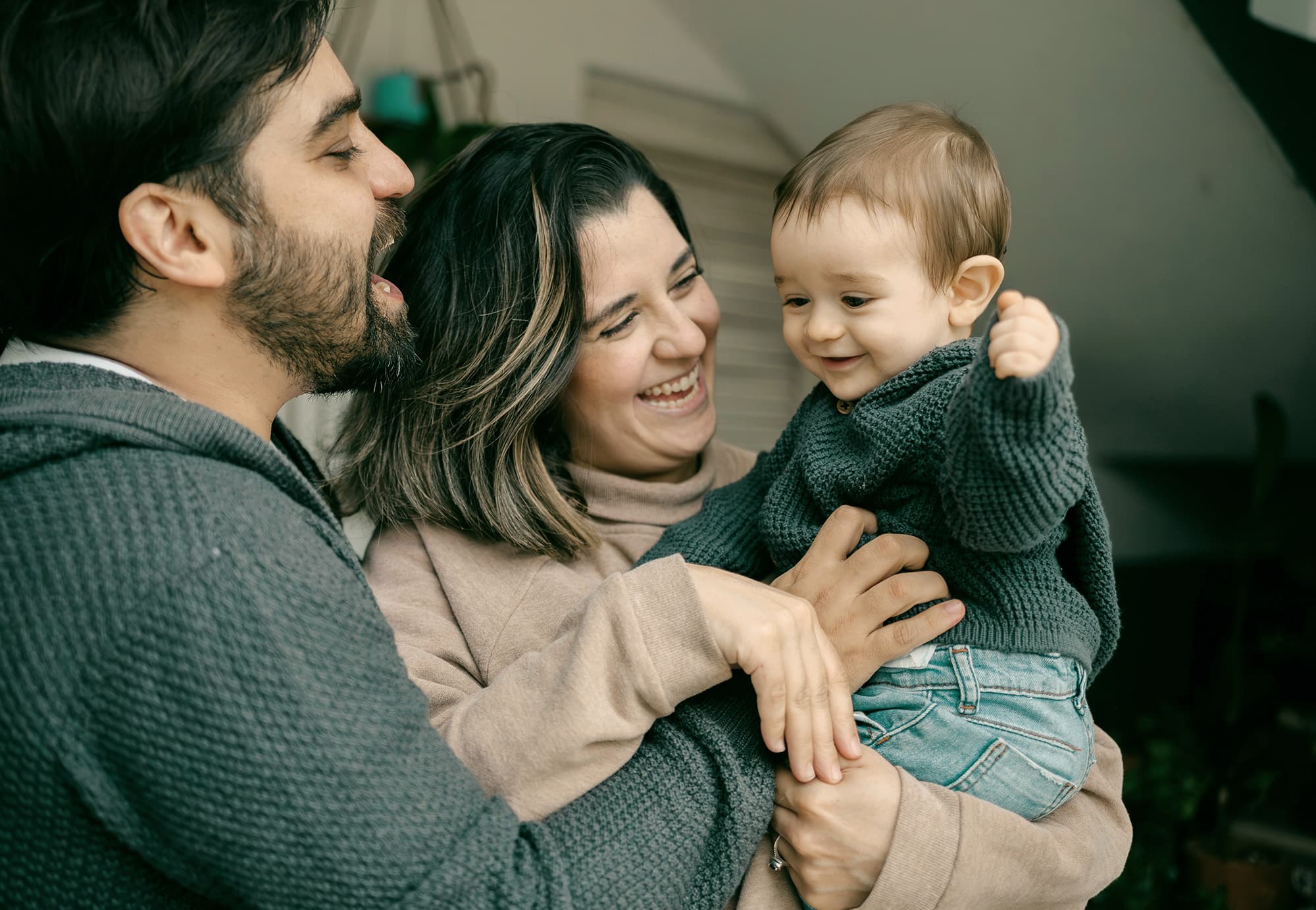 A joyful family with a bearded man on the far left in a grey shirt tickling a laughing baby’s stomach, the mother in the center smiling happily at the baby while holding her, as the baby, dressed in a greenish-grey knitted top and jeans, raises a hand and giggles.