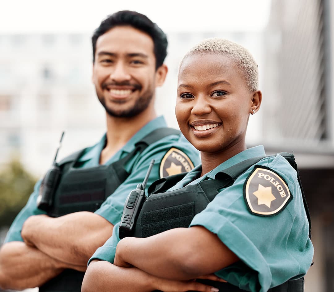 A woman police officer in a teal uniform and black vest with a walkie-talkie stands confidently, smiling, while a man in the same uniform is seen smiling in the blurred background.