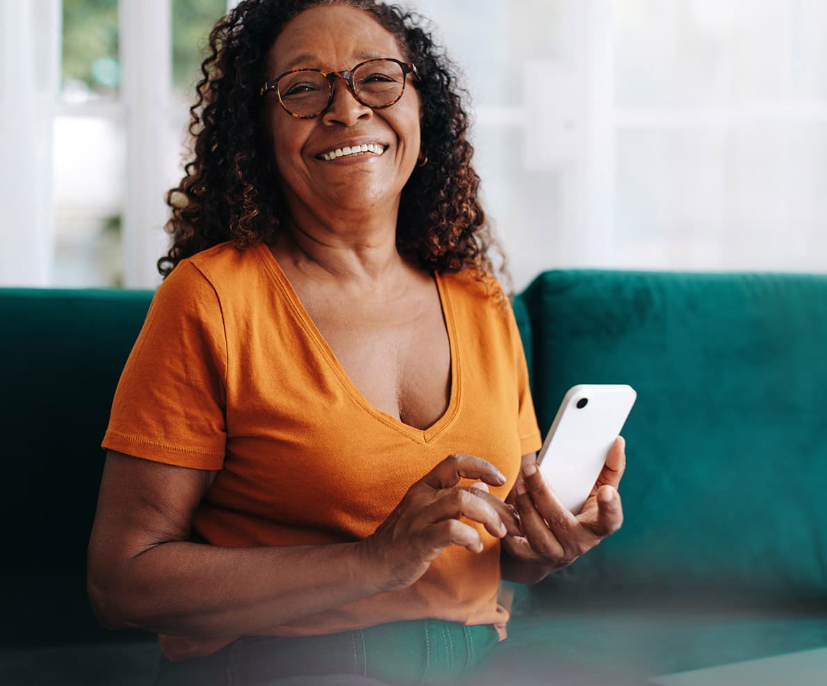 A smiling woman sits on a dark teal couch with an iPad beside her, wearing an orange T-shirt and holding an iPhone.