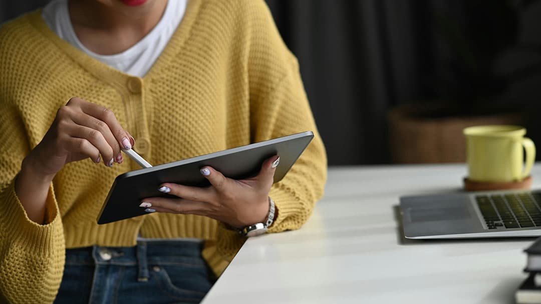 A close-up of a person holding an iPad and drawing on it with a pen; the screen is not visible. They are wearing a yellow knitted cardigan over a white shirt and jeans, sitting sideways in front of a table with a computer and a yellow mug.