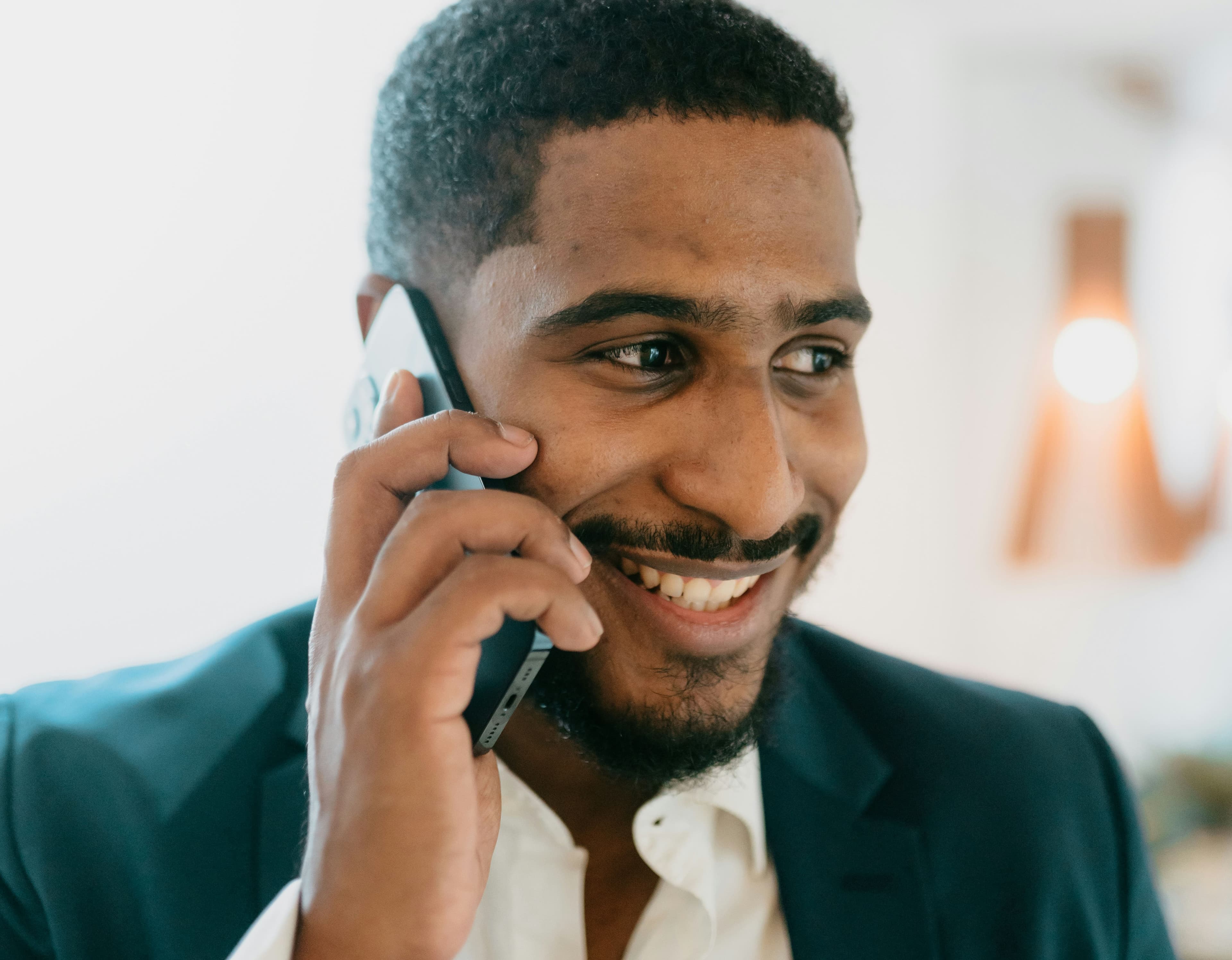 A close-up of a bearded man smiling while talking on the phone, wearing a dark teal suit over a white shirt.