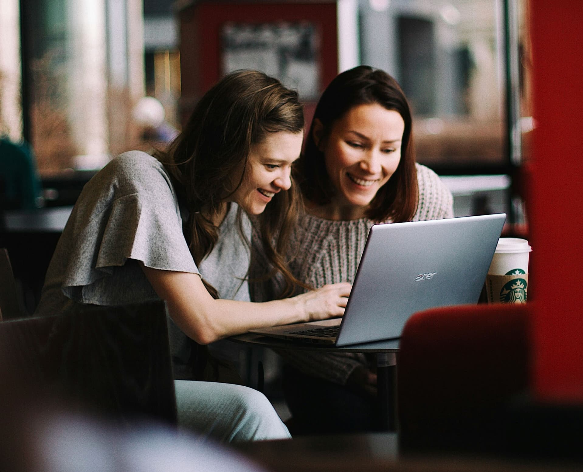 Two women sitting together at a café table, smiling as they look at a computer, with red wallpaper in the background.