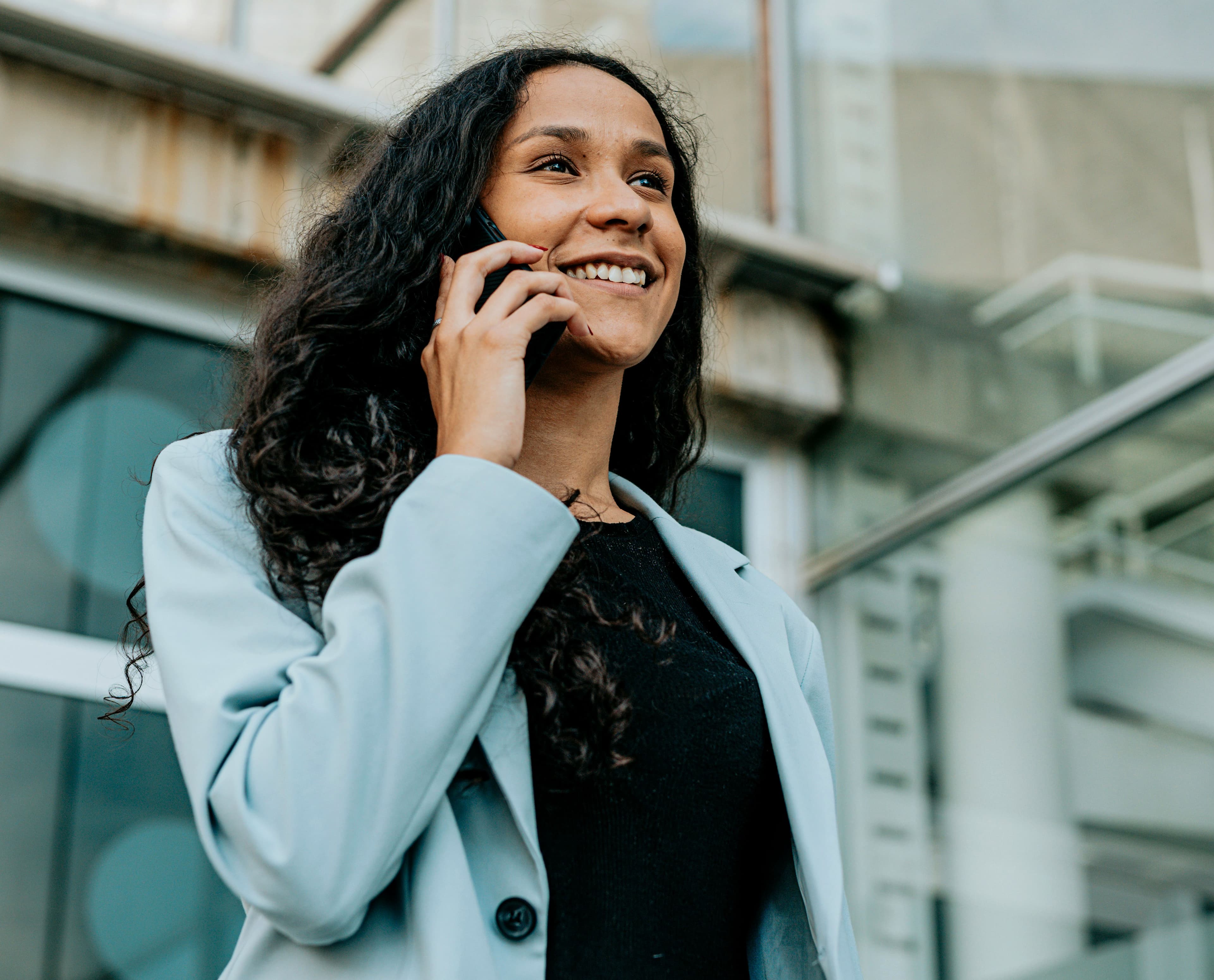 A woman with her hair down stands outside, smiling while on the phone and looking ahead hopefully, dressed in a light blue suit and a black top.
