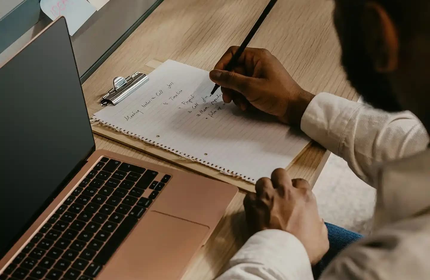 A close-up of a man's hand writing in a notebook on a table, with a laptop beside him, capturing a focused moment of note-taking.