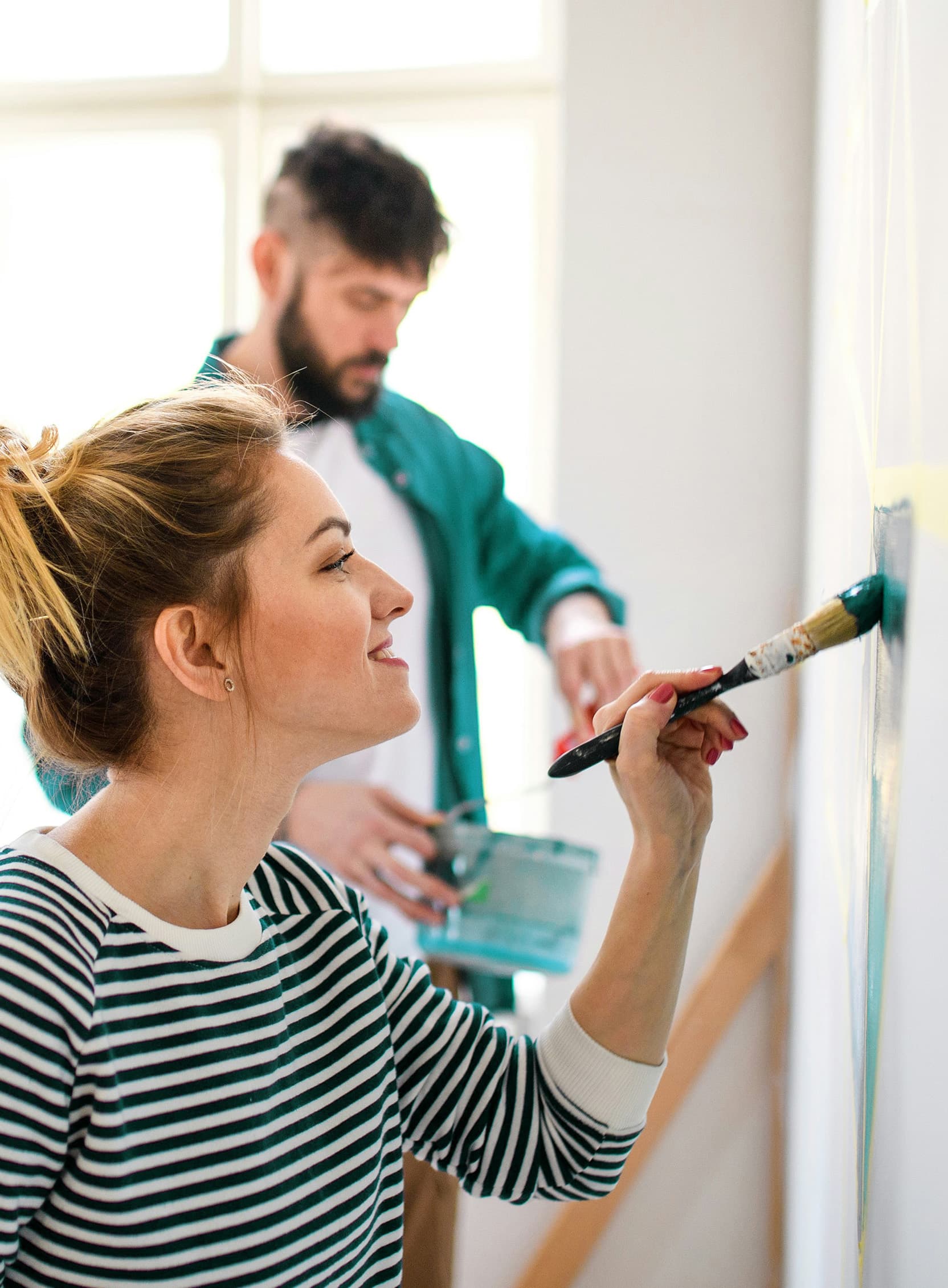 A close-up of a blonde woman painting a white wall with teal paint, wearing a striped shirt, while in the blurred background, a man in a teal shirt layered over a white shirt is visible.