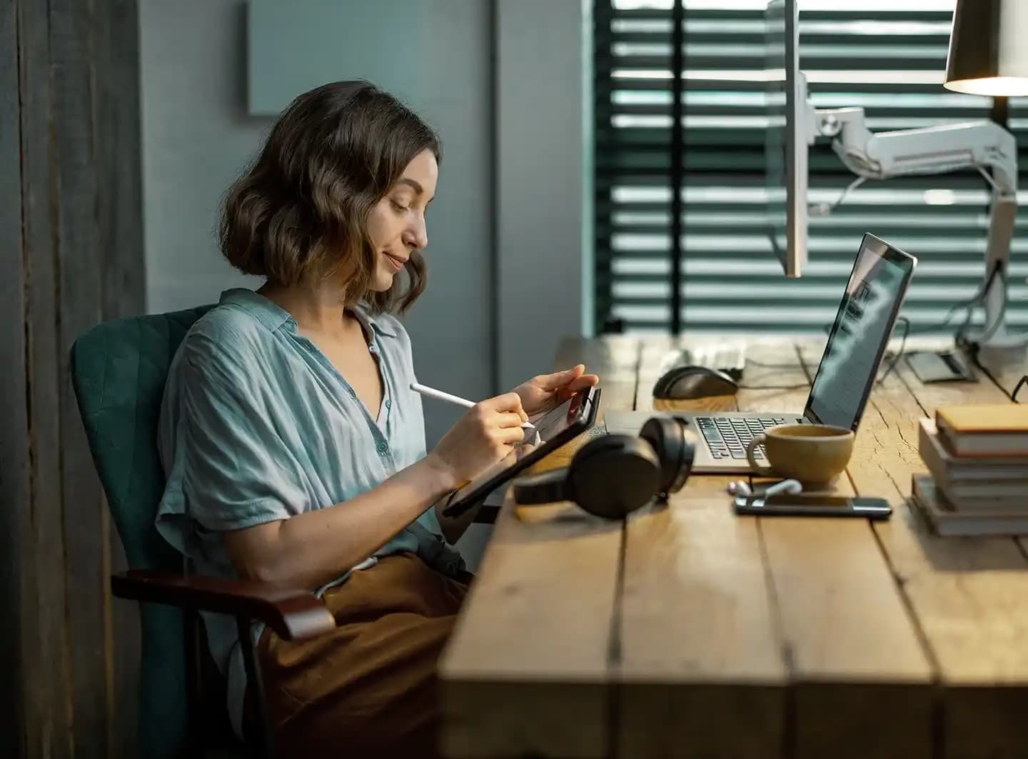 A woman with a bob cut sitting at a table with a laptop and monitor, holding an iPad and drawing on it using an iPad pen.