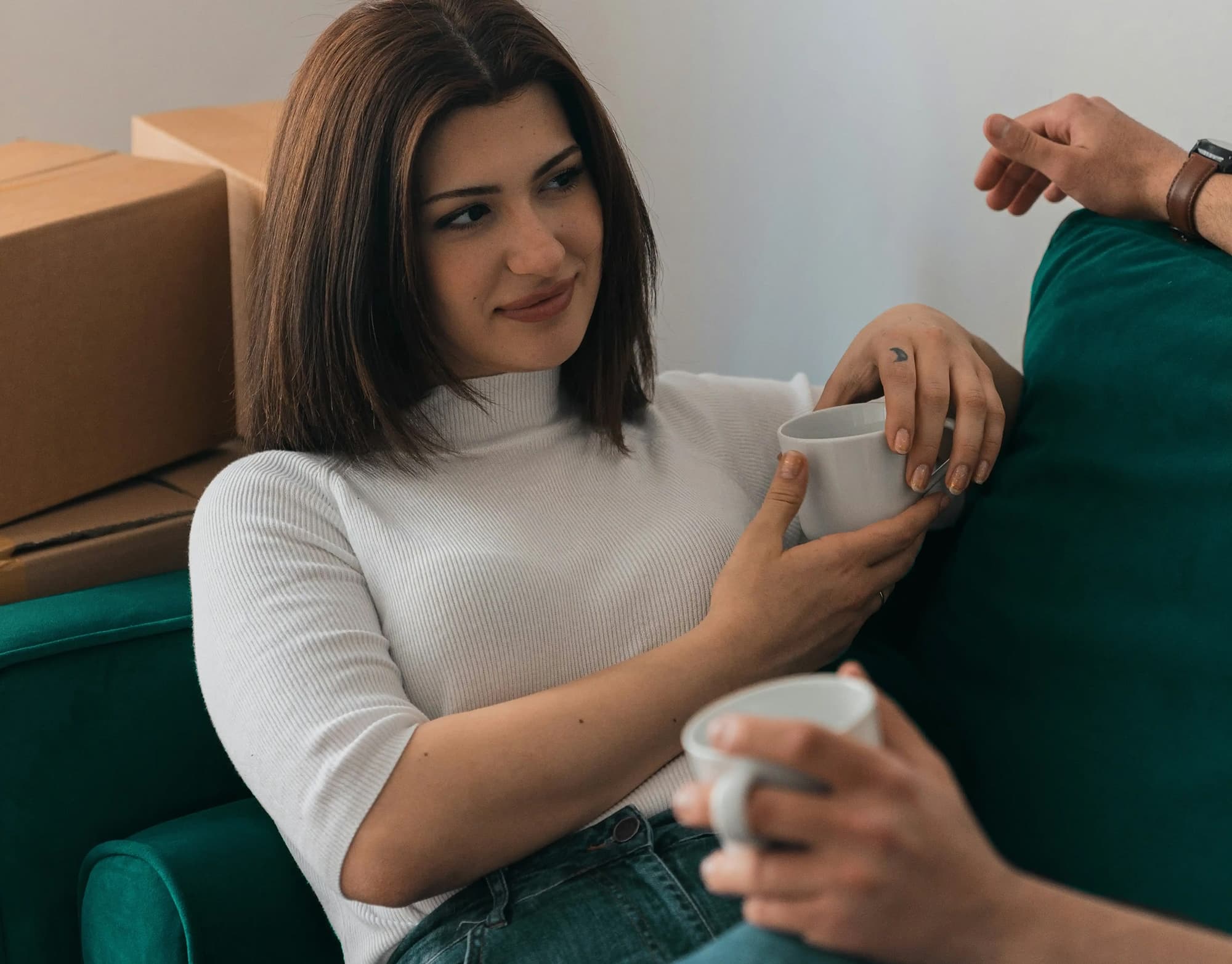 A woman sitting comfortably on a couch, holding a white coffee mug and smiling as she listens to another person.