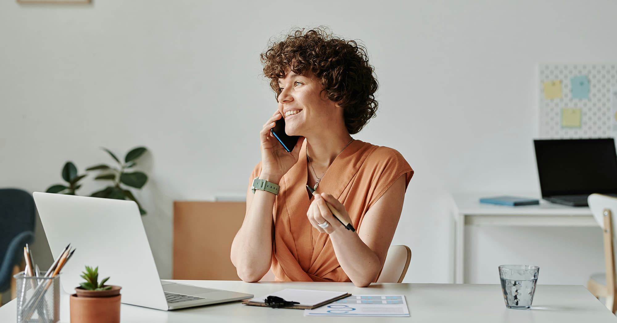 A woman with short curly brown hair sits at her white office desk, smiling while holding a phone to her ear and looking to the side; she wears an orange shirt and a green smartwatch, with a pen in her other hand, and in front of her are an open laptop, documents, a glass of water, a small potted plant, and a pencil holder filled with pencils.